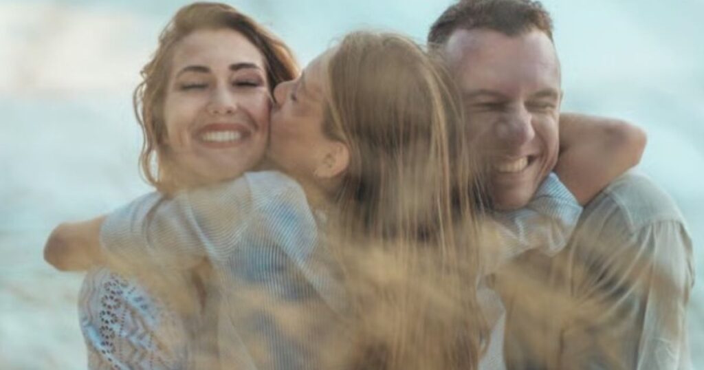 A smiling couple outdoors by the water, with a child kissing the woman on the cheek